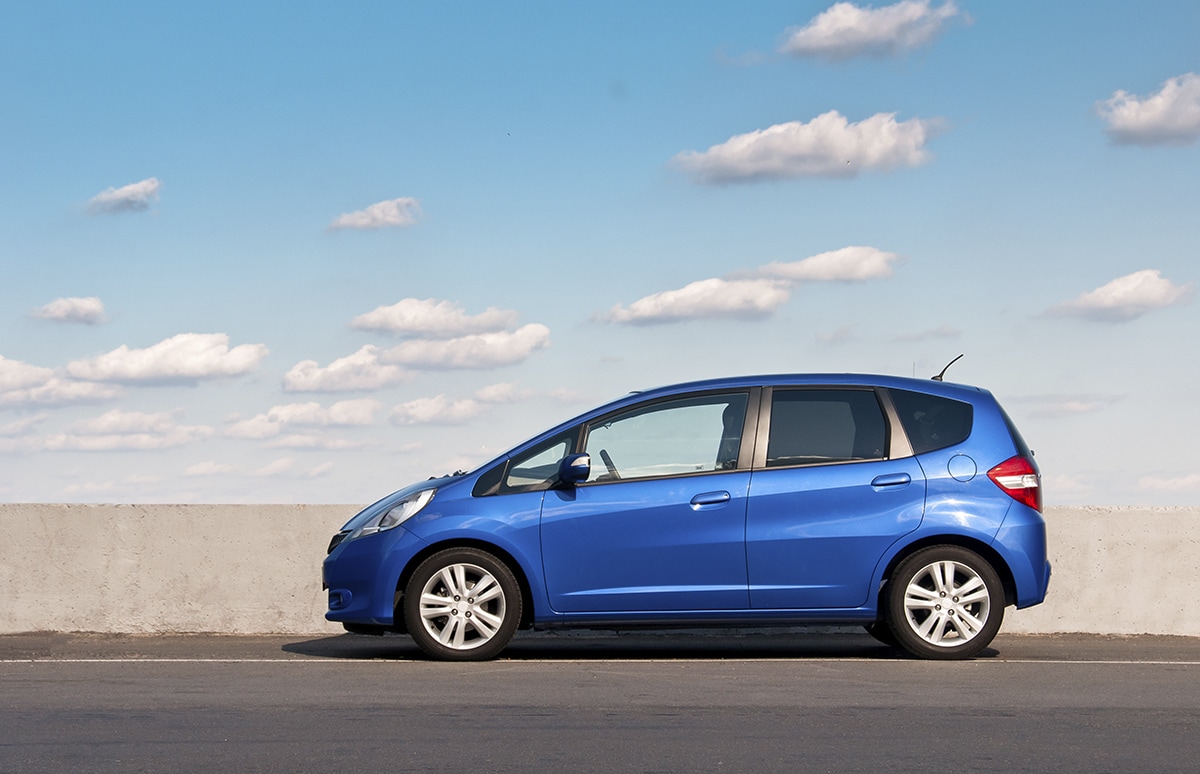Coche compacto azul visto de perfil, estacionado junto a un muro de hormigón bajo un cielo despejado con nubes dispersas