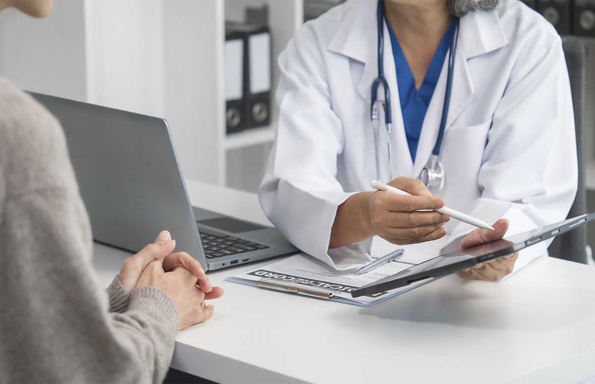 Profesional sanitario mostrando información en una tableta durante una consulta, con documentos y un portátil sobre la mesa