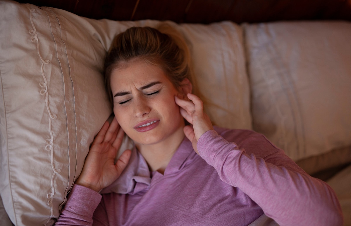 Persona recostada en una cama sujetándose ambos lados de la mandíbula, una imagen que ilustra molestias mandibulares asociadas al bruxismo o al rechinamiento dental durante el descanso