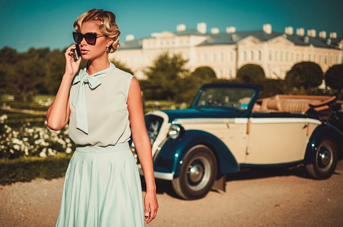 Mujer vestida con estilo retro hablando por teléfono mientras un elegante coche clásico descapotable permanece estacionado al fondo