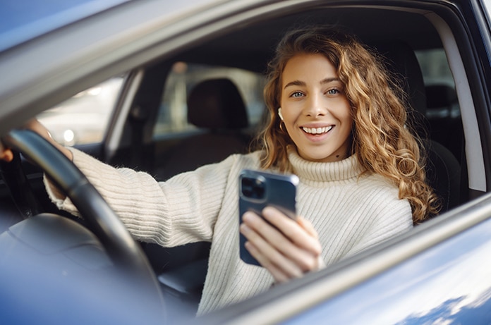 Conductora dentro de su vehículo consultando su teléfono móvil mientras mantiene la otra mano en el volante