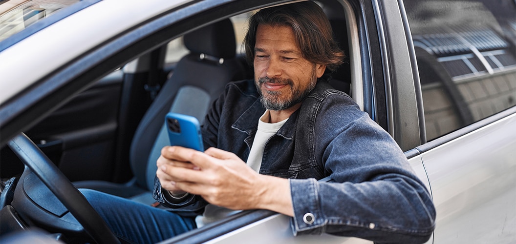Persona sentada en el asiento del conductor de un vehículo, utilizando un teléfono móvil con la ventanilla bajada en un entorno urbano