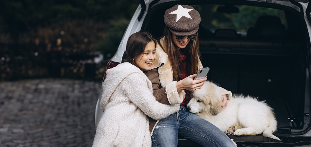Madre e hija con su mascota apolladas en el maletero abierto de su coche sonrientes mirando un móvil