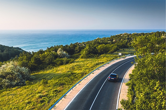 Vehículo circulando por una carretera costera rodeada de vegetación con vistas al mar y el horizonte despejado