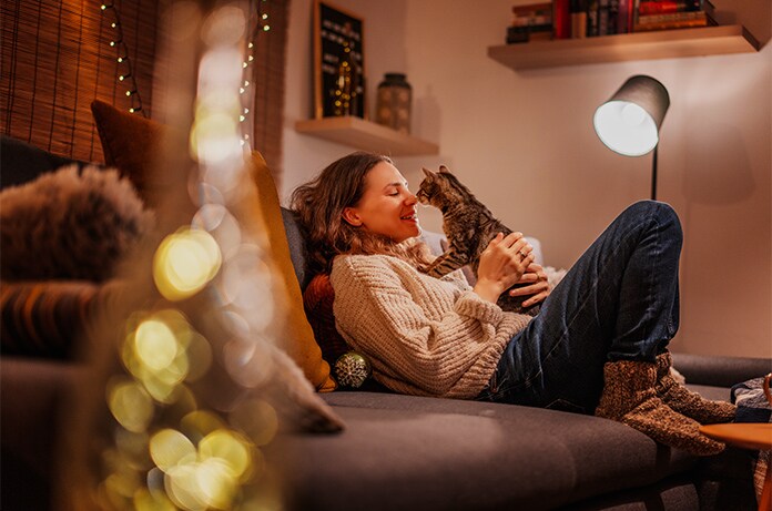 Figura recostada en un sofá en un salón cálido y decorado, sosteniendo un gato mientras descansa entre luces ambientales