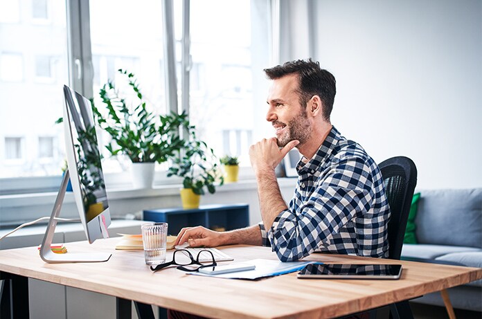 Figura sentada en un escritorio moderno mirando un monitor, rodeada de documentos, gafas y una bebida en vaso transparente