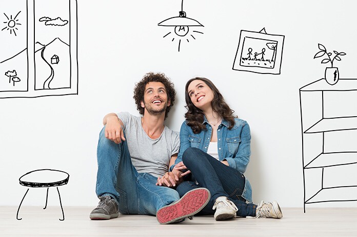 Pareja sentada en el suelo frente a una pared decorada con ilustraciones de un hogar, en una escena simbólica de vida doméstica