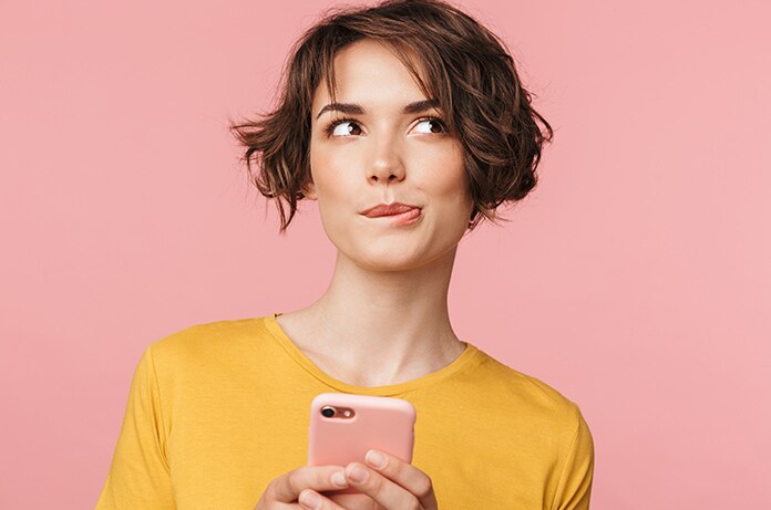Persona sosteniendo un teléfono móvil frente a un fondo liso de color rosa, con vestimenta en tonos cálidos y composición minimalista