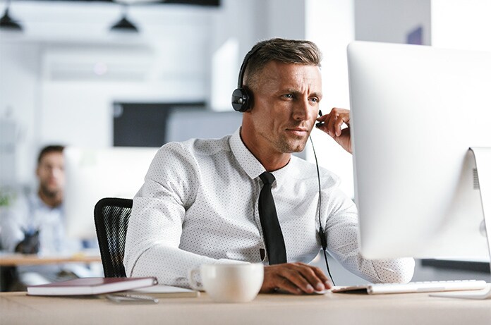 Persona trabajando en una oficina moderna frente a un ordenador de sobremesa, utilizando auriculares y material de oficina sobre la mesa