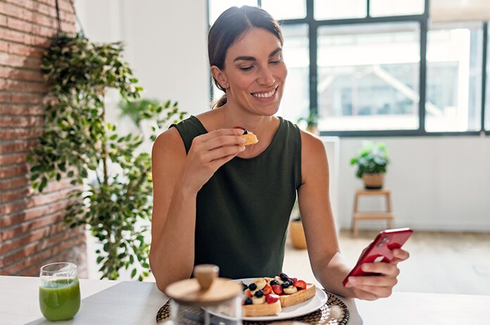 Persona sentada a una mesa durante una comida ligera, utilizando un teléfono móvil junto a un plato con alimentos y una bebida, en un espacio luminoso con plantas
