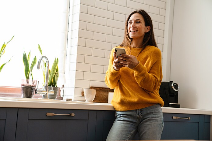 Persona apoyada en una encimera de cocina moderna, utilizando un teléfono móvil junto a plantas y pequeños electrodomésticos