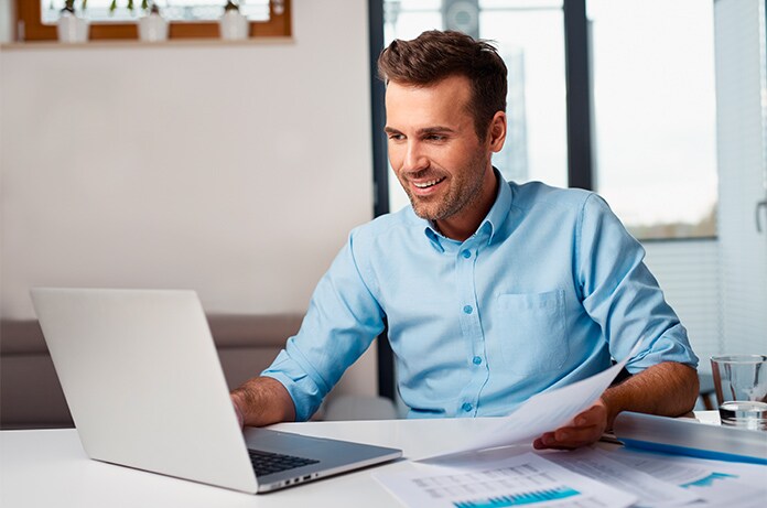Persona trabajando con un ordenador portátil mientras revisa documentos impresos en una mesa de trabajo iluminada