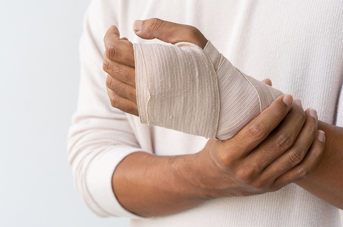 Primer plano de una mano vendada, sujetada con la otra mano, sobre un fondo claro y neutro