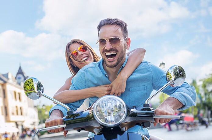 Dos personas abrazándose mientras circulan en un ciclomotor por una zona peatonal con edificios al fondo