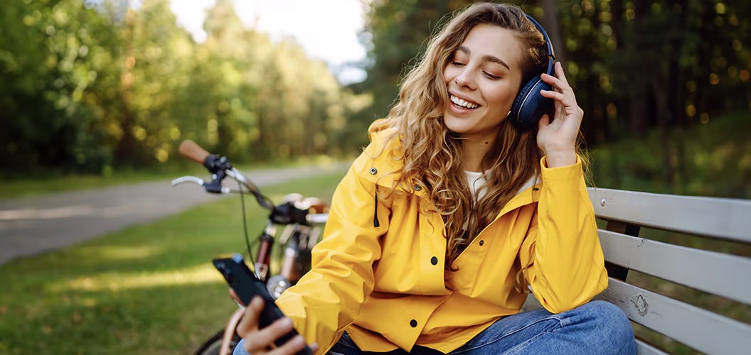 Persona con auriculares sentada en un banco junto a una bicicleta mientras consulta un teléfono móvil en un entorno natural