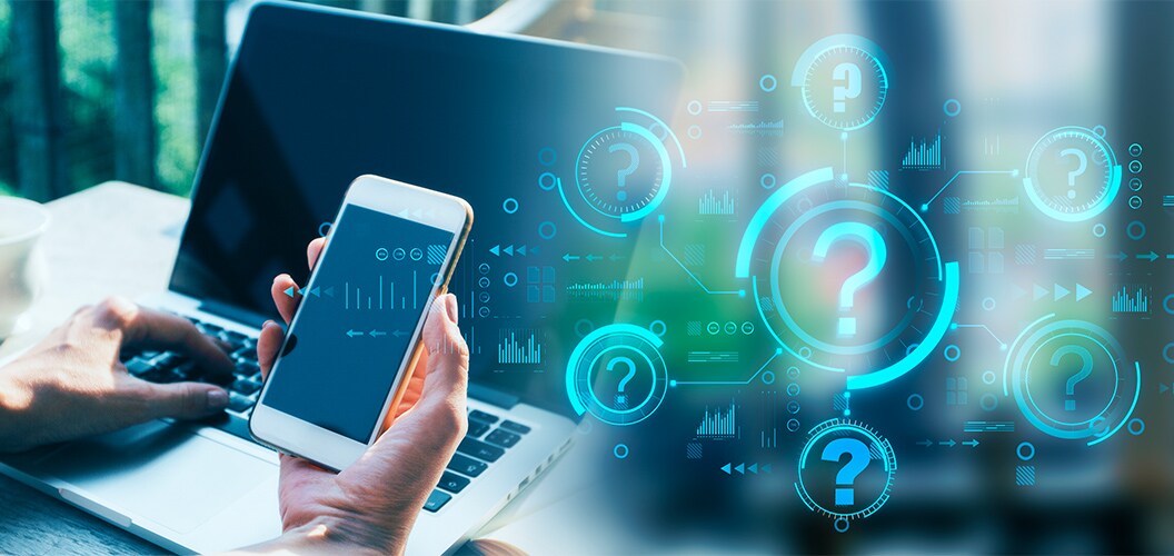 Mano sosteniendo un teléfono móvil frente a un ordenador portátil, rodeado de iconos digitales con signos de interrogación